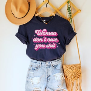 A navy blue unisex crew neck t-shirt with the words "women don't owe you shit" in retro pink text with retro pink stars