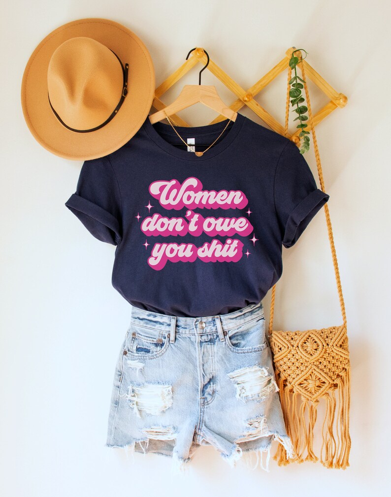 A navy blue unisex crew neck t-shirt with the words "women don't owe you shit" in retro pink text with retro pink stars