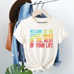 A natural (light beige) crew neck short sleeve unisex t-shirt that says "welcome to the coolest summer of the rest of your life" with a blue, green, yellow and red heat map in the text