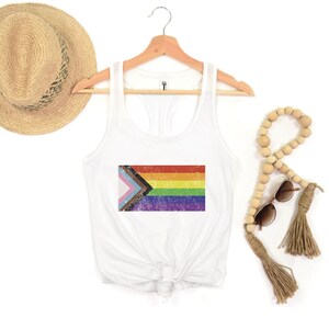 May include: White tank top with a rainbow pride flag printed on the front. The flag has a distressed, vintage look. The tank top is on a hanger.