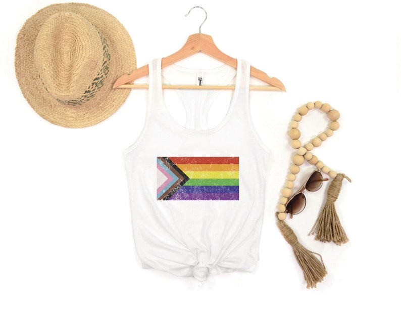 May include: White tank top with a rainbow pride flag printed on the front. The flag has a distressed, vintage look. The tank top is on a hanger.