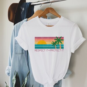 May include: White t-shirt with a retro sunset and palm trees graphic. The text "Respect It. Protect It" is printed below the graphic.