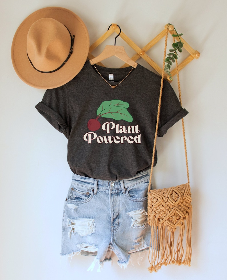 May include: A dark gray t-shirt with a cherry and leaves graphic and the text "Plant Powered" in a retro font. The shirt is on a hanger with a straw hat and a woven bag.