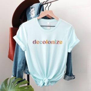 以下が含まれることがあります： 明るいブルーのTシャツに、カラフルな文字で「脱植民地化」と印刷されています。