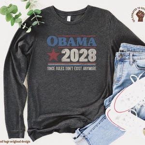 May include: A dark charcoal gray long-sleeve t-shirt featuring a distressed graphic print that reads OBAMA 2028 with a red star and the text SINCE RULES DON'T EXIST ANYMORE. The shirt is paired with light blue denim jeans and white canvas sneakers.