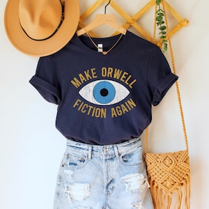 A navy blue unisex crew neck Bella Canvas brand t-shirt that says "MAKE ORWELL FICTION AGAIN" in orange text with a black outline, around an image of a blue eye. The graphic has a distressed vintaged look.