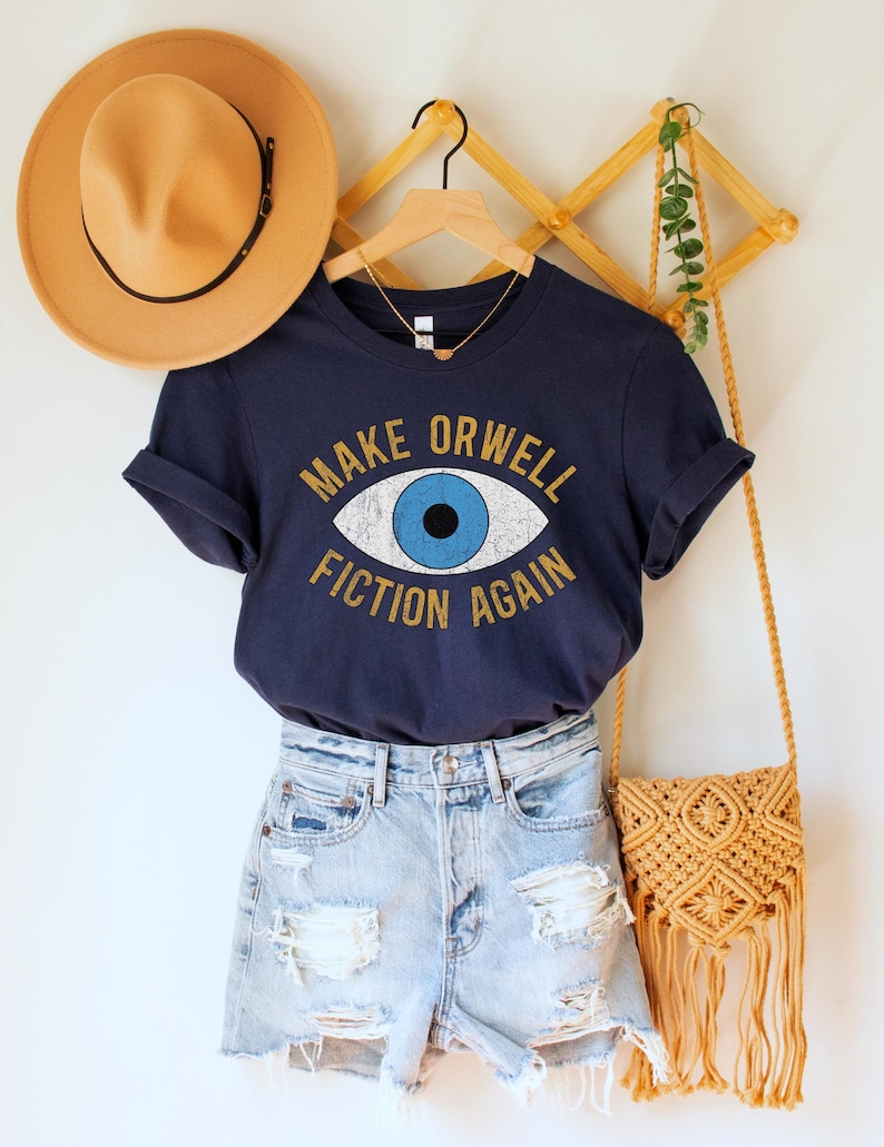 A navy blue unisex crew neck Bella Canvas brand t-shirt that says "MAKE ORWELL FICTION AGAIN" in orange text with a black outline, around an image of a blue eye. The graphic has a distressed vintaged look.