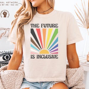 May include: Cream-colored t-shirt with the text "THE FUTURE IS INCLUSIVE" in a bold, black font. The design features a colorful sunburst with rainbow-colored rays. The shirt is a casual, unisex style.