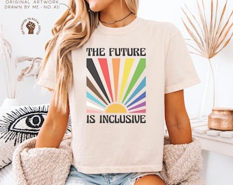 May include: Cream-colored t-shirt with the text "THE FUTURE IS INCLUSIVE" in a bold, black font. The design features a colorful sunburst with rainbow-colored rays. The shirt is a casual, unisex style.