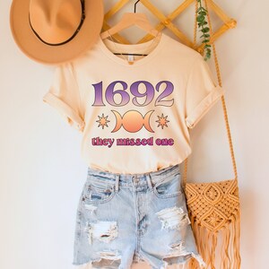 Puede incluir: Una camiseta beige claro con un estampado gr&aacute;fico del n&uacute;mero 1692, un s&iacute;mbolo de triple luna y el texto "they missed one".