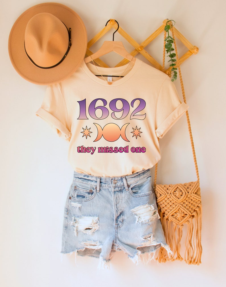 Puede incluir: Una camiseta beige claro con un estampado gr&aacute;fico del n&uacute;mero 1692, un s&iacute;mbolo de triple luna y el texto "they missed one".
