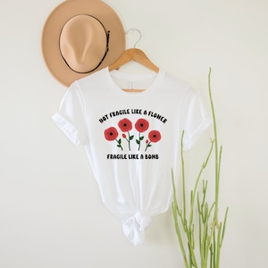 May include: White t-shirt with a graphic of red flowers and the text "Not Fragile Like A Flower Fragile Like A Bomb".