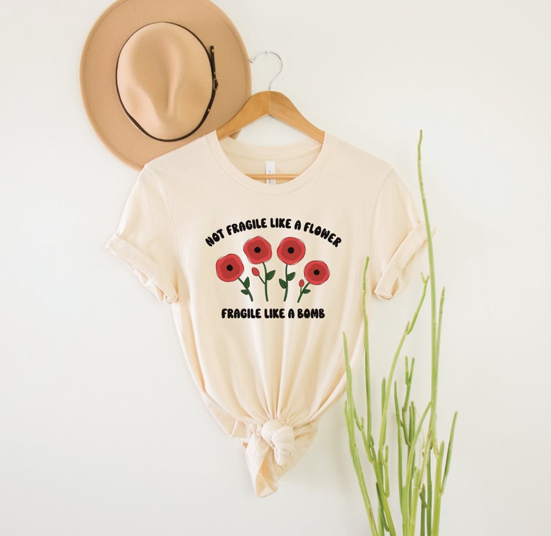 May include: A beige t-shirt with a graphic print of red flowers and the text "Not Fragile Like A Flower, Fragile Like A Bomb".