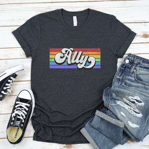 May include: A dark grey t-shirt with a rainbow stripe design and the word "Ally" in a retro font.