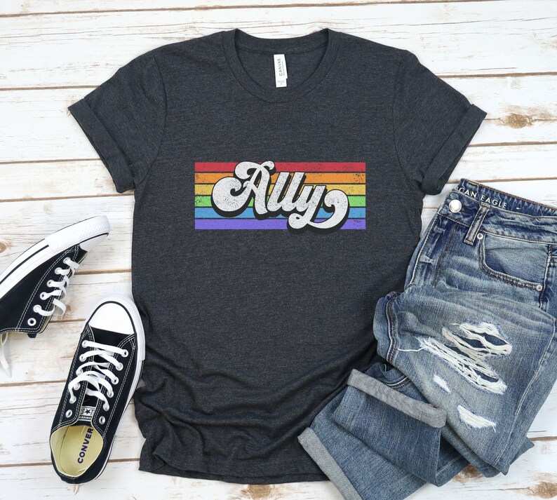 May include: A dark grey t-shirt with a rainbow stripe design and the word "Ally" in a retro font.