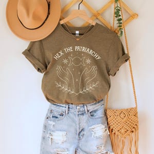A heather olive green/brown unisex crew neck short sleeve Bella+Canvas t-shirt that says "hex the patriarchy" in a white uppercase distressed font, above a witchy white graphic with leaves, hands, stars, and a three moon symbol