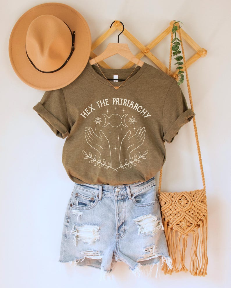A heather olive green/brown unisex crew neck short sleeve Bella+Canvas t-shirt that says "hex the patriarchy" in a white uppercase distressed font, above a witchy white graphic with leaves, hands, stars, and a three moon symbol