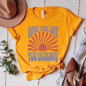 A gold (dark yellow) unisex short sleeve crew neck t-shirt that says "have the day you deserve" on the front in distressed blue retro capitalized font around a distressed image of a red wavy retro sunset
