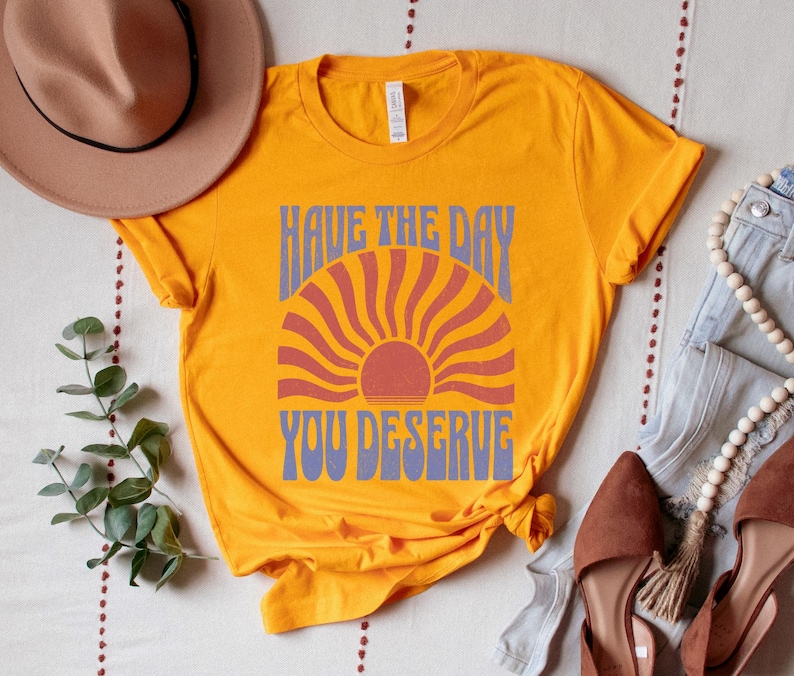 A gold (dark yellow) unisex short sleeve crew neck t-shirt that says "have the day you deserve" on the front in distressed blue retro capitalized font around a distressed image of a red wavy retro sunset