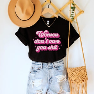 A black unisex crew neck t-shirt with the words "women don't owe you shit" in retro pink text with retro pink stars
