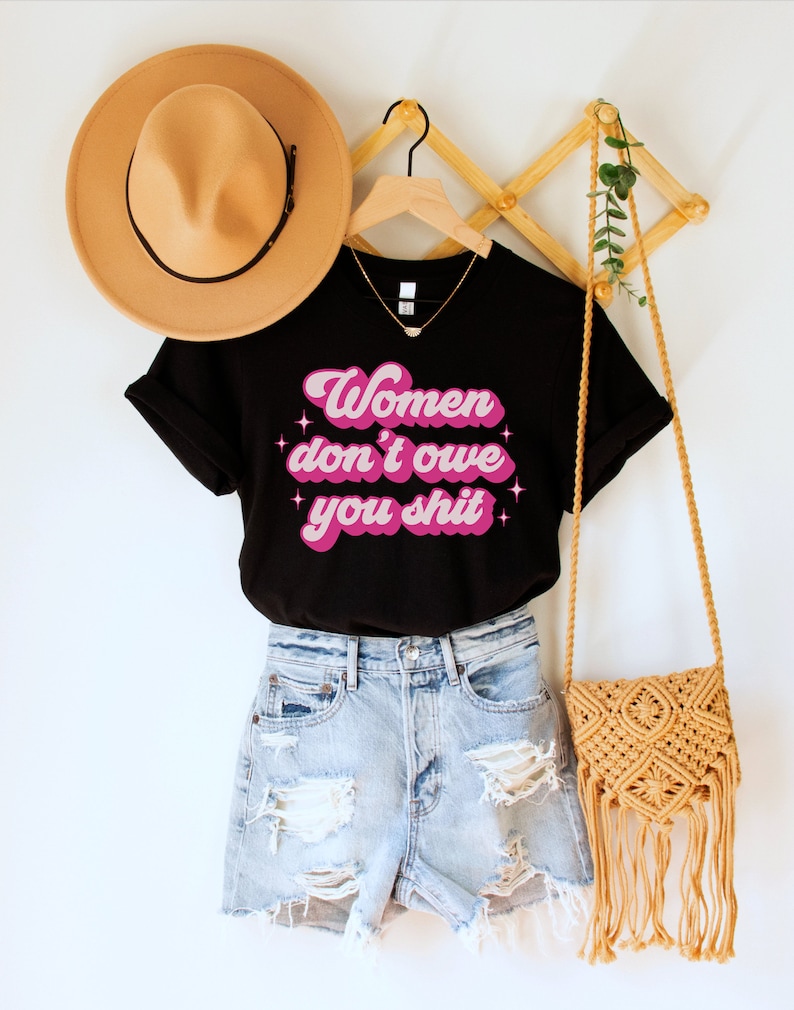 A black unisex crew neck t-shirt with the words "women don't owe you shit" in retro pink text with retro pink stars