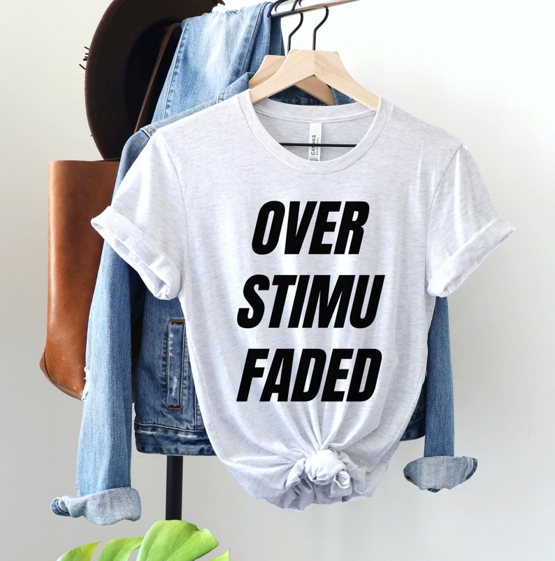 May include: Light gray t-shirt with the words "OVER STIMU FADED" printed in bold black letters. The shirt is hanging on a wooden hanger with a denim jacket and a brown leather bag in the background.