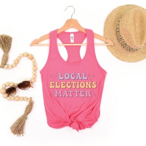 A hot pink women’s racerback tank top that says "local elections matter" in purple, yellow, and pink retro text with retro stars