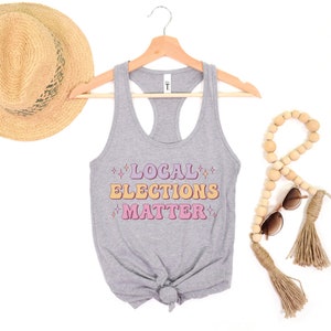 A heather medium grey green women’s racerback tank top that says "local elections matter" in purple, yellow, and pink retro text with retro stars