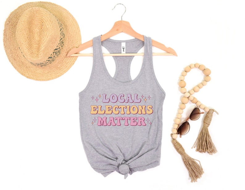 A heather medium grey green women’s racerback tank top that says "local elections matter" in purple, yellow, and pink retro text with retro stars