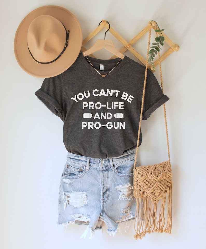 Peut inclure: Un t-shirt gris fonc&eacute; avec l'inscription blanche "YOU CAN'T BE PRO-LIFE AND PRO-GUN". Il est associ&eacute; &agrave; un short en jean, un chapeau beige et un sac en macram&eacute;. Un collier dor&eacute; est visible.