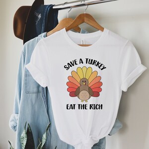K&ouml;nnte beinhalten: Wei&szlig;es T-Shirt mit einem Cartoon-Truthahn-Design und dem Text "Save a Turkey Eat the Rich". Der Truthahn hat einen braunen K&ouml;rper und einen bunten F&auml;cher aus Federn in Gelb-, Orange- und Rott&ouml;nen.