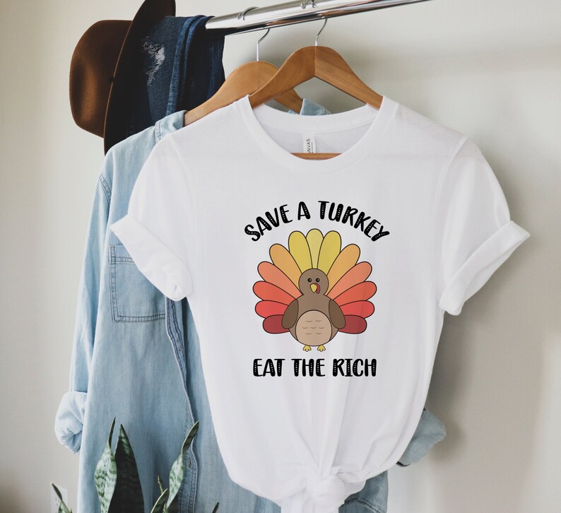 K&ouml;nnte beinhalten: Wei&szlig;es T-Shirt mit einem Cartoon-Truthahn-Design und dem Text "Save a Turkey Eat the Rich". Der Truthahn hat einen braunen K&ouml;rper und einen bunten F&auml;cher aus Federn in Gelb-, Orange- und Rott&ouml;nen.