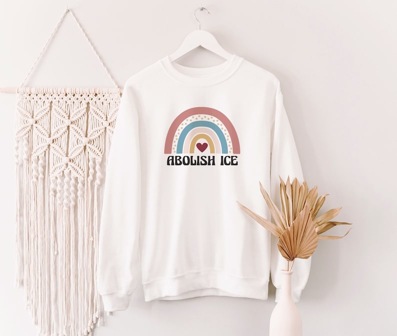 May include: White sweatshirt with a rainbow graphic and the text "Abolish ICE" in black letters. The rainbow is made up of pink, blue, yellow, and orange stripes with a red heart in the center.