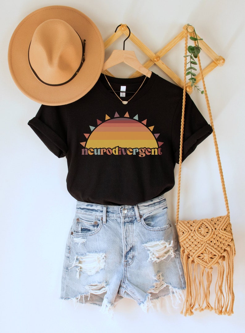 May include: Black t-shirt with a retro sunset graphic and the text "neurodivergent". The shirt is styled with denim shorts and a brown woven bag.
