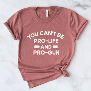 Peut inclure: Un t-shirt de couleur rose poussi&eacute;reux avec l'inscription blanche "YOU CAN'T BE PRO-LIFE AND PRO-GUN" et des graphiques de balles. Le t-shirt est pli&eacute; et les manches sont retrouss&eacute;es, pour un style d&eacute;contract&eacute;.