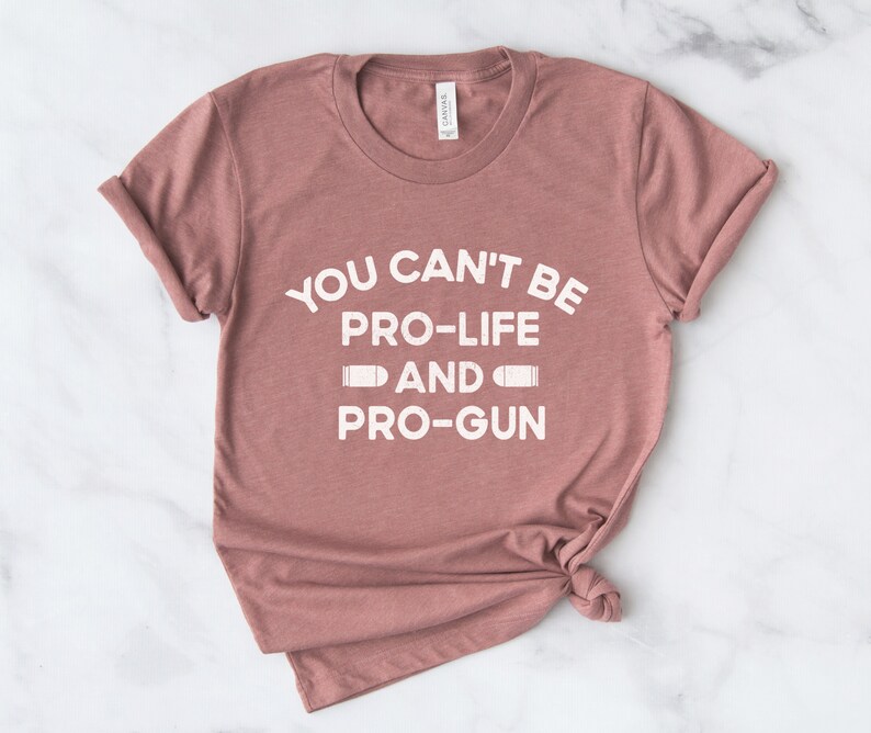 Peut inclure: Un t-shirt de couleur rose poussi&eacute;reux avec l'inscription blanche "YOU CAN'T BE PRO-LIFE AND PRO-GUN" et des graphiques de balles. Le t-shirt est pli&eacute; et les manches sont retrouss&eacute;es, pour un style d&eacute;contract&eacute;.