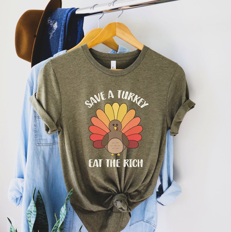 K&ouml;nnte beinhalten: Olivgr&uuml;nes T-Shirt mit einem Truthahn-Motiv und dem Text "Save a Turkey Eat the Rich". Der Truthahn hat einen bunten F&auml;cher aus Federn in Gelb-, Orange- und Rott&ouml;nen. Das T-Shirt h&auml;ngt an einem Holzb&uuml;gel.