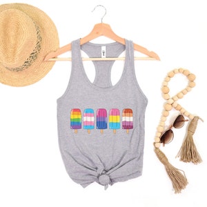 Pu&ograve; includere: Un top grigio senza maniche con un design di ghiaccioli con i colori dell'arcobaleno, la bandiera transgender e la bandiera bisessuale. Il top &egrave; su un appendiabiti con un cappello di paglia e perline di legno sullo sfondo.