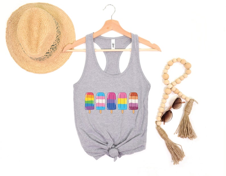 Pu&ograve; includere: Un top grigio senza maniche con un design di ghiaccioli con i colori dell'arcobaleno, la bandiera transgender e la bandiera bisessuale. Il top &egrave; su un appendiabiti con un cappello di paglia e perline di legno sullo sfondo.