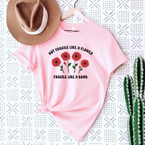 May include: Pink t-shirt with a graphic of red flowers and the text "Not Fragile Like A Flower Fragile Like A Bomb".