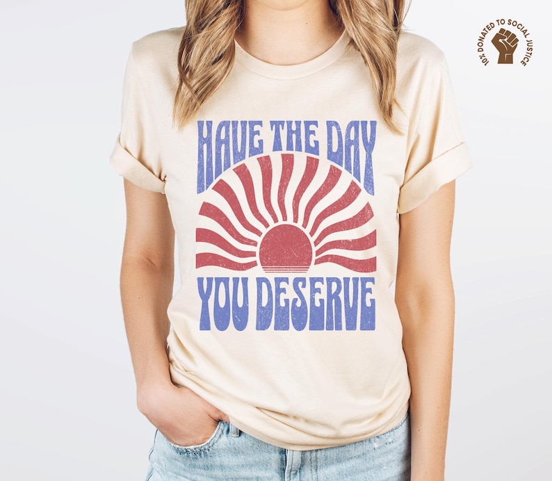 A natural (light beige) unisex short sleeve crew neck t-shirt that says "have the day you deserve" on the front in distressed blue retro capitalized font around a distressed image of a red wavy retro sunset