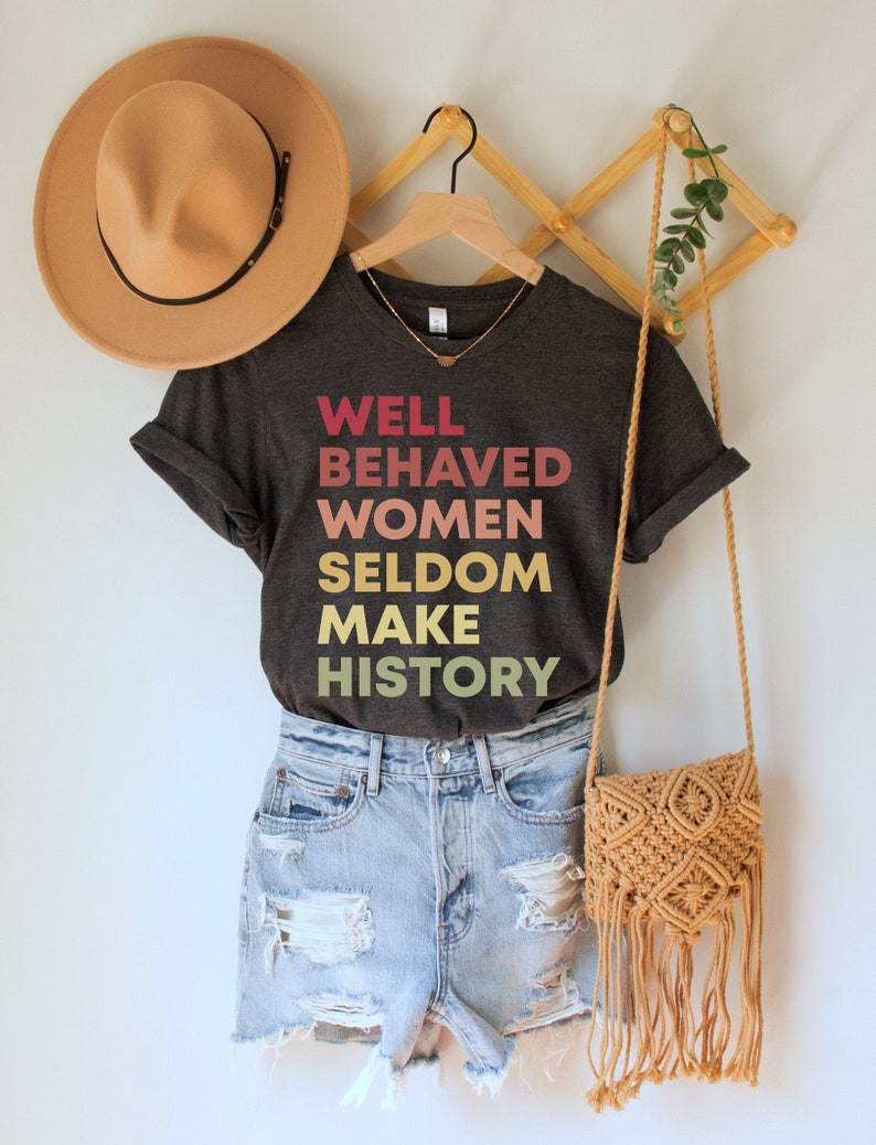 May include: A dark gray t-shirt with the text "WELL BEHAVED WOMEN SELDOM MAKE HISTORY" printed in a gradient of pink, orange, and yellow.