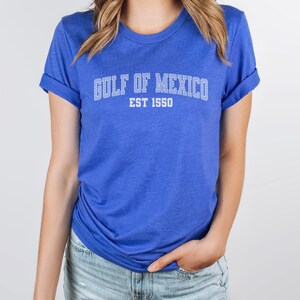 A heather true royal blue unisex crew neck short sleeve Bella+Canvas brand t-shirt that says "GULF OF MEXICO" on the chest in white outline college style font with white stripes inside the letters. The text is curved over smaller solid white letters in the same font that say "EST 1550". The graphic has a distressed/vintage look