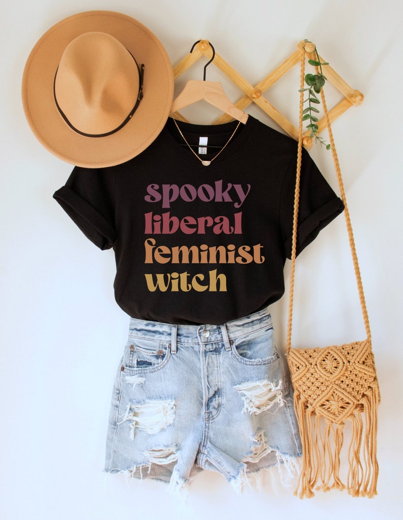 A black unisex crew neck t-shirt with the phrase "spooky liberal feminist witch" in purple, red, orange, and yellow text