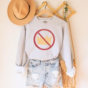 An oatmeal (light grey) heather unisex crew neck long sleeve fleece-lined sweatshirt with an image of a yellow crown inside a red circle with a slash over it, meaning "no kings". The image has a vintage/distressed look.