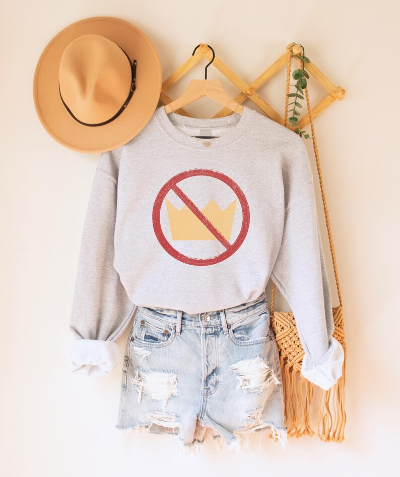 An oatmeal (light grey) heather unisex crew neck long sleeve fleece-lined sweatshirt with an image of a yellow crown inside a red circle with a slash over it, meaning "no kings". The image has a vintage/distressed look.