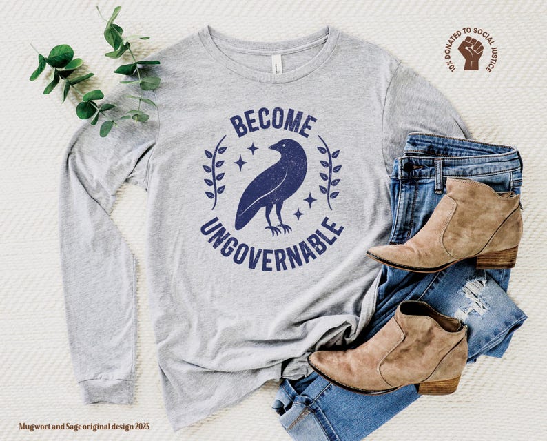 An athletic (medium grey) heather unisex long sleeve crew neck t-shirt that says  "BECOME UNGOVERNABLE" on the chest in block text around an image of a crow, leaves, and retro stars. The whole graphic and text are dark blue and have a distressed/vintage look.