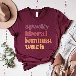 A maroon unisex crew neck t-shirt with the phrase "spooky liberal feminist witch" in purple, red, orange, and yellow text