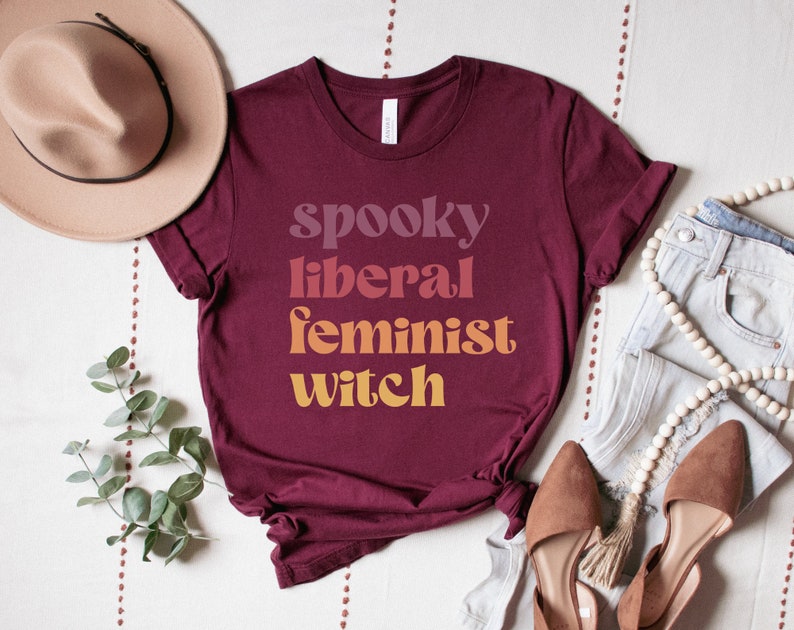A maroon unisex crew neck t-shirt with the phrase "spooky liberal feminist witch" in purple, red, orange, and yellow text