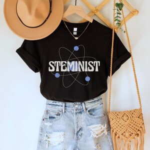 May include: Black t-shirt with a white atom graphic and the text "STEMINIST" in white letters. The shirt is on a hanger with a brown hat and a woven bag.